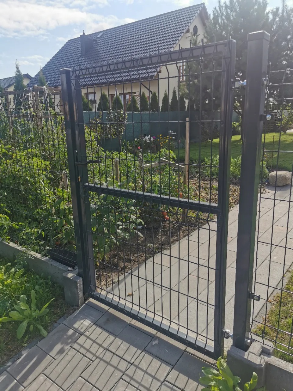 Otwarta, metalowa furtka panelowa w kolorze grafitowym, prowadząca do ogrodu z widocznymi grządkami warzywnymi i domem jednorodzinnym w tle.