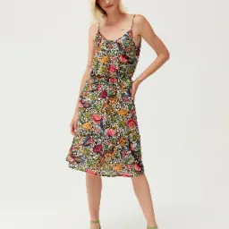 Modelka prezentuje letnią sukienkę na ramiączkach z motywem kwiatowo-ptasim na białym tle, uzupełnioną o zielone sandałki na obcasie.