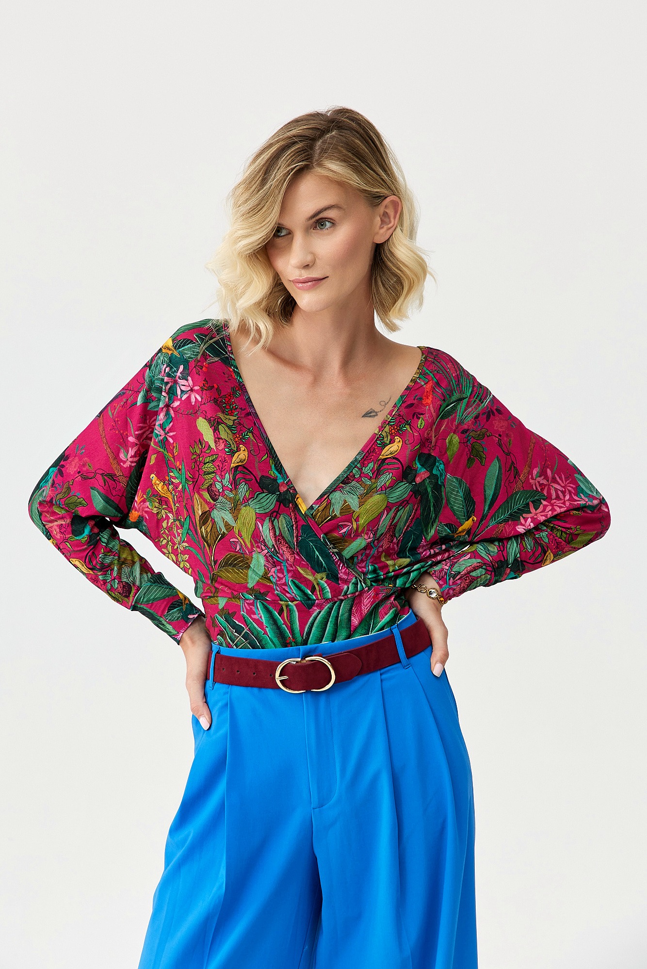 Kobieta pozuje w bluzce z tropikalnym wzorem i szerokich, niebieskich spodniach z bordowym paskiem na białym tle.