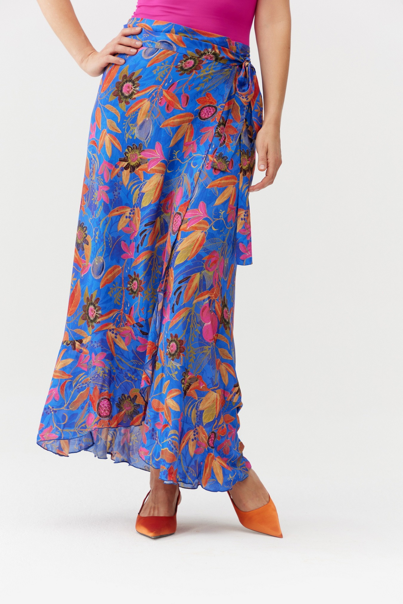 Kobieta prezentuje długą, zwiewną spódnicę w odcieniach błękitu z wzorem kwiatowym w kolorach różu, pomarańczy i brązu, uzupełnioną o pomarańczowe szpilki.