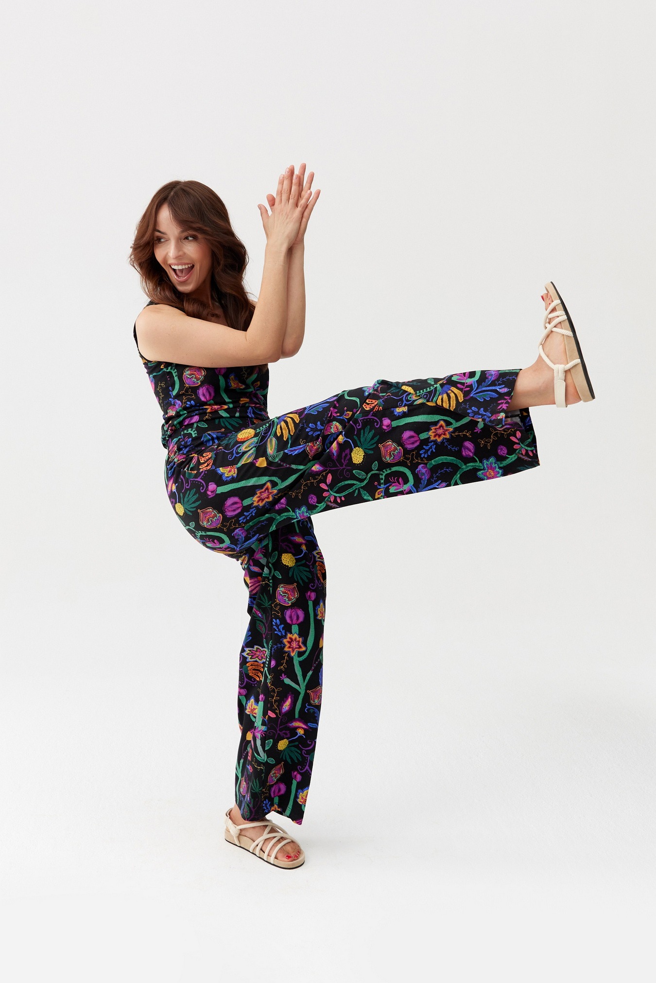 Uśmiechnięta kobieta w czarnym, wzorzystym kombinezonie i sandałach na platformie, pozująca na białym tle z uniesioną nogą.
