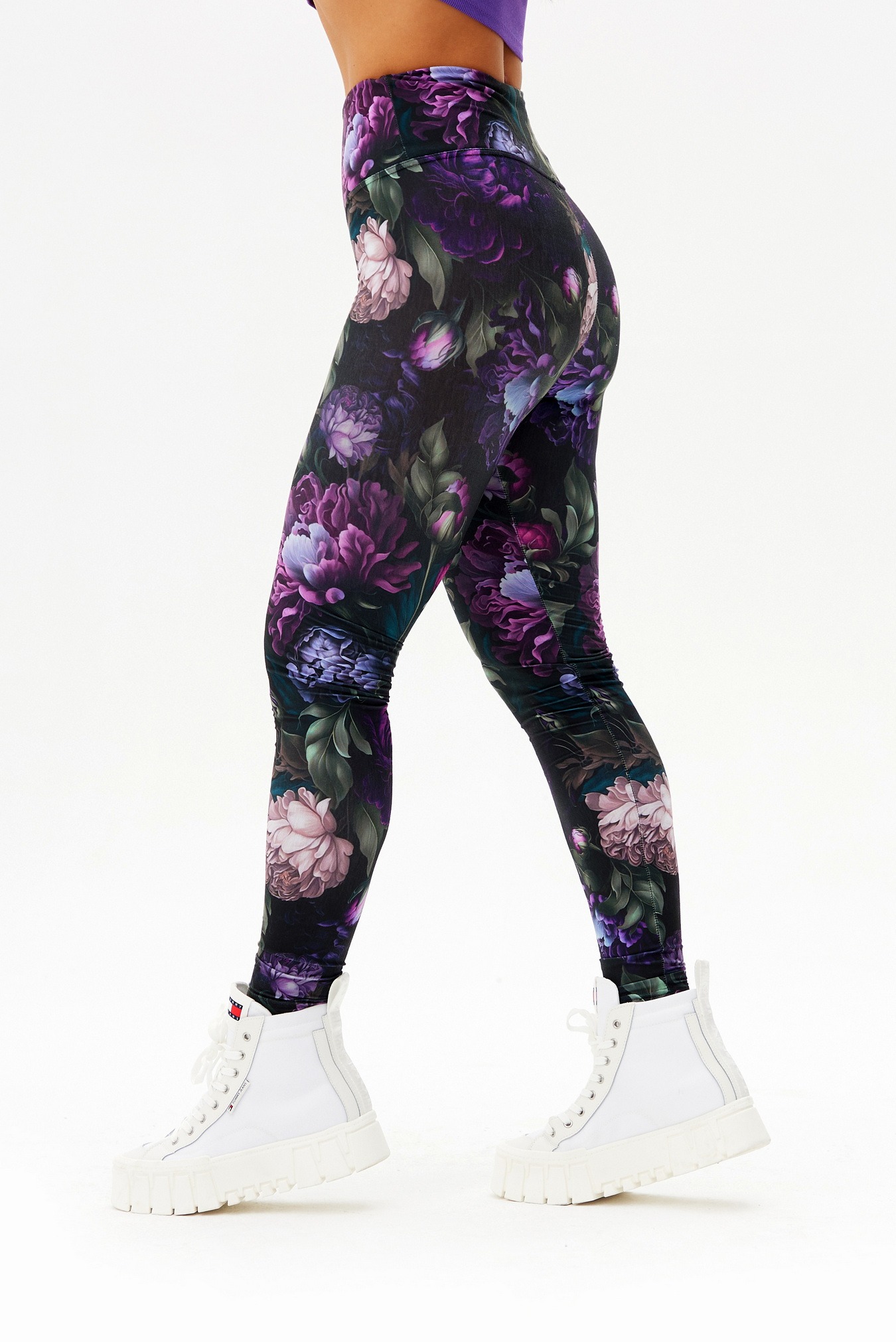 Kobieta w legginsach z motywem ciemnych, fioletowych kwiatów i białych butach sportowych na platformie, na białym tle.