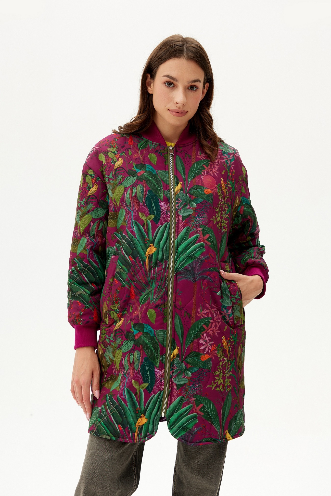 Kobieta prezentuje długą, pikowaną kurtkę bomberkę w tropikalne wzory z motywami ptaków i roślin na białym tle.