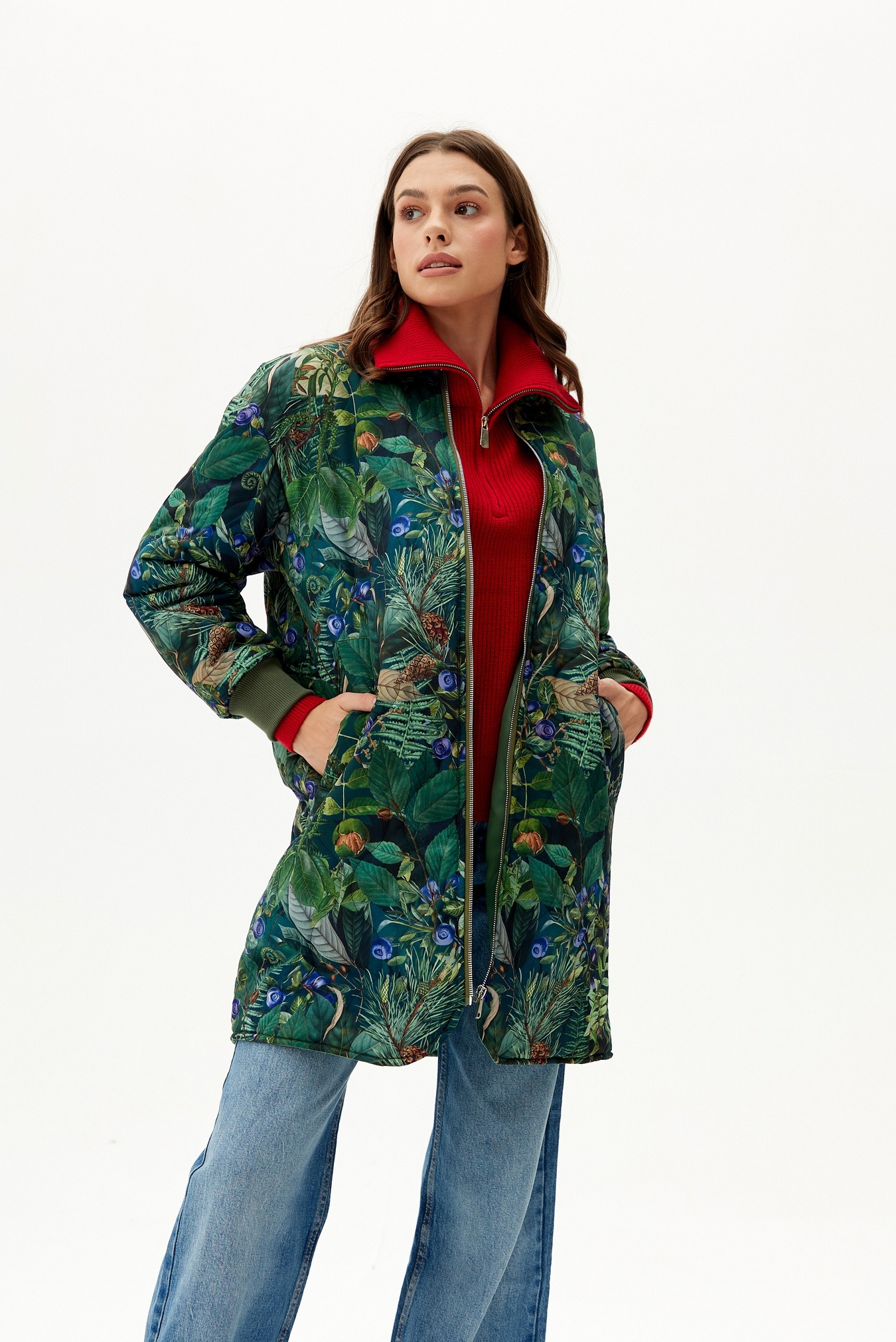 Kobieta w dżinsach i długiej kurtce bomber z wzorem liści, igieł sosnowych i jagód na białym tle.