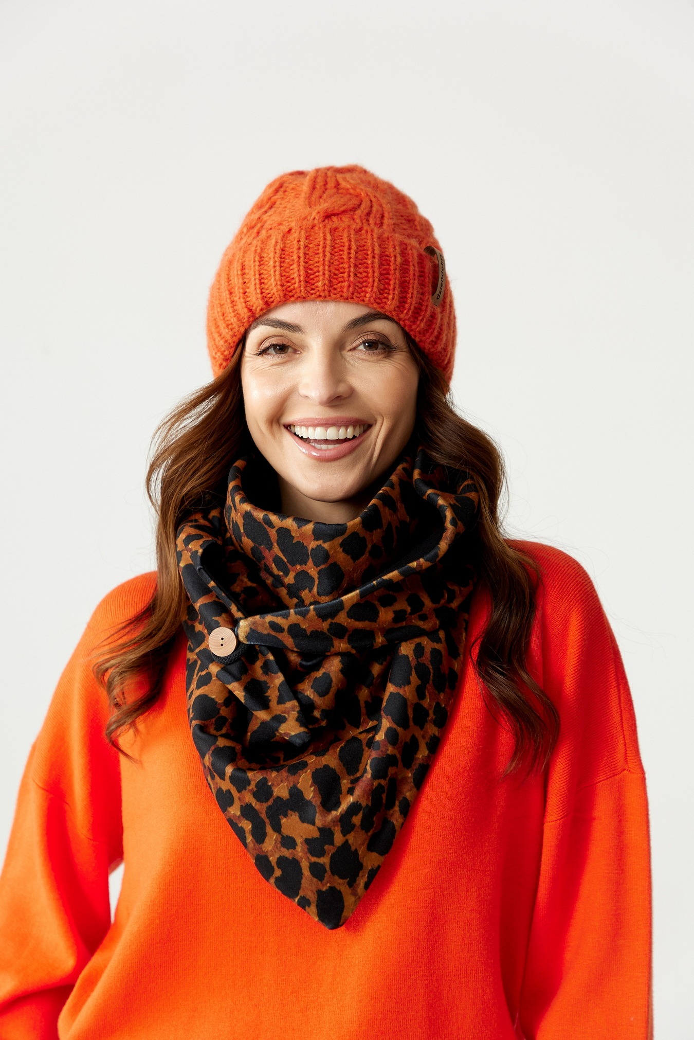 Uśmiechnięta kobieta w pomarańczowym swetrze i czapce, z apaszką w panterkę zapinaną na drewniany guzik.