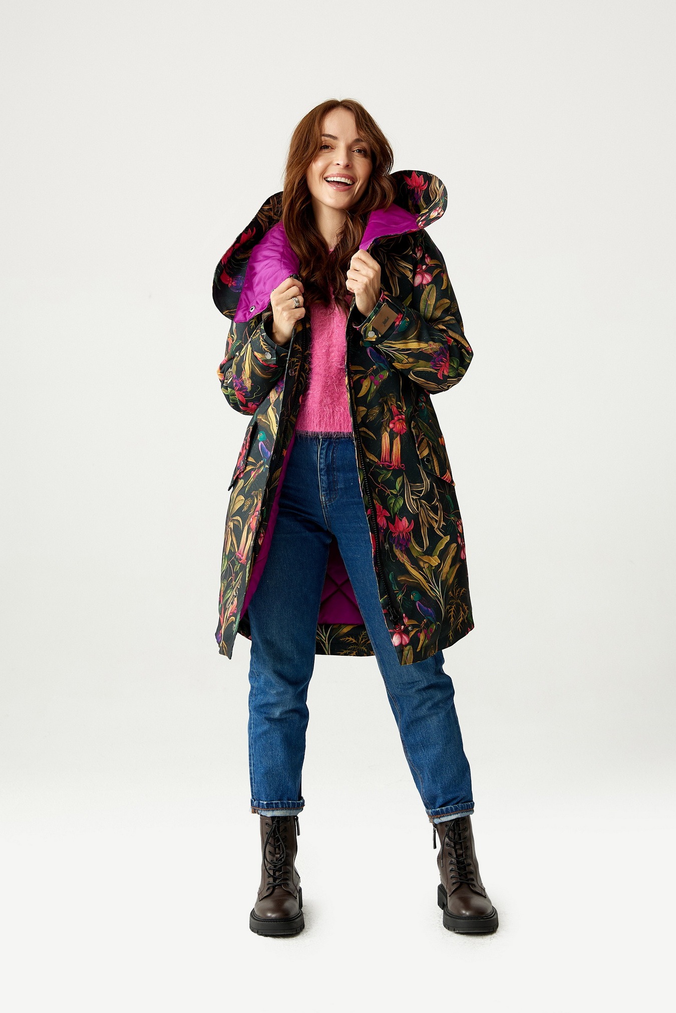 Uśmiechnięta kobieta w długim płaszczu z motywem tropikalnych kwiatów i ptaków, z fioletową podszewką, ubrana w różowy sweter i dżinsy, pozuje na białym tle.