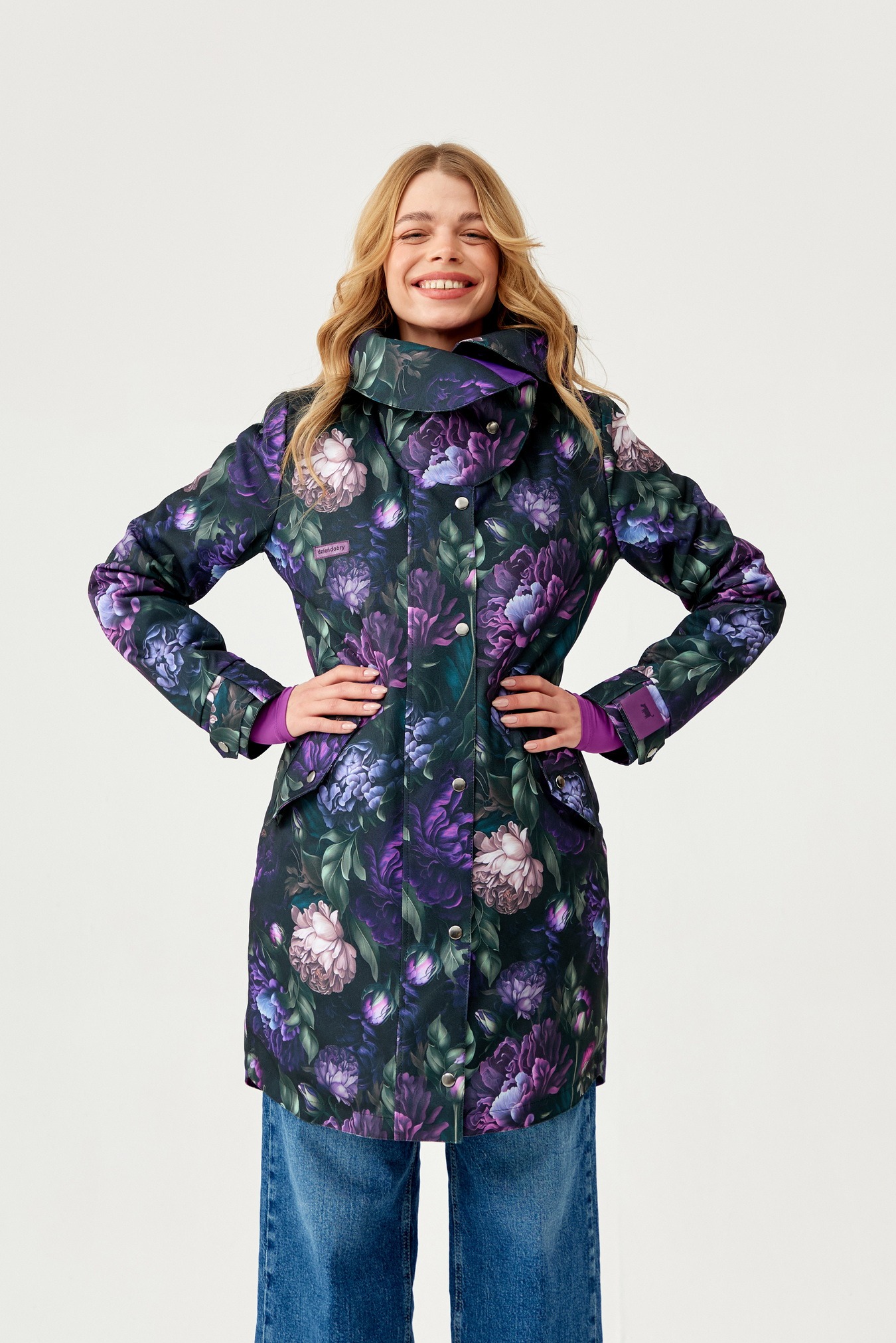 Uśmiechnięta blondynka w płaszczu z motywem fioletowych kwiatów, prezentuje odzież damską w Łodzi na białym tle.