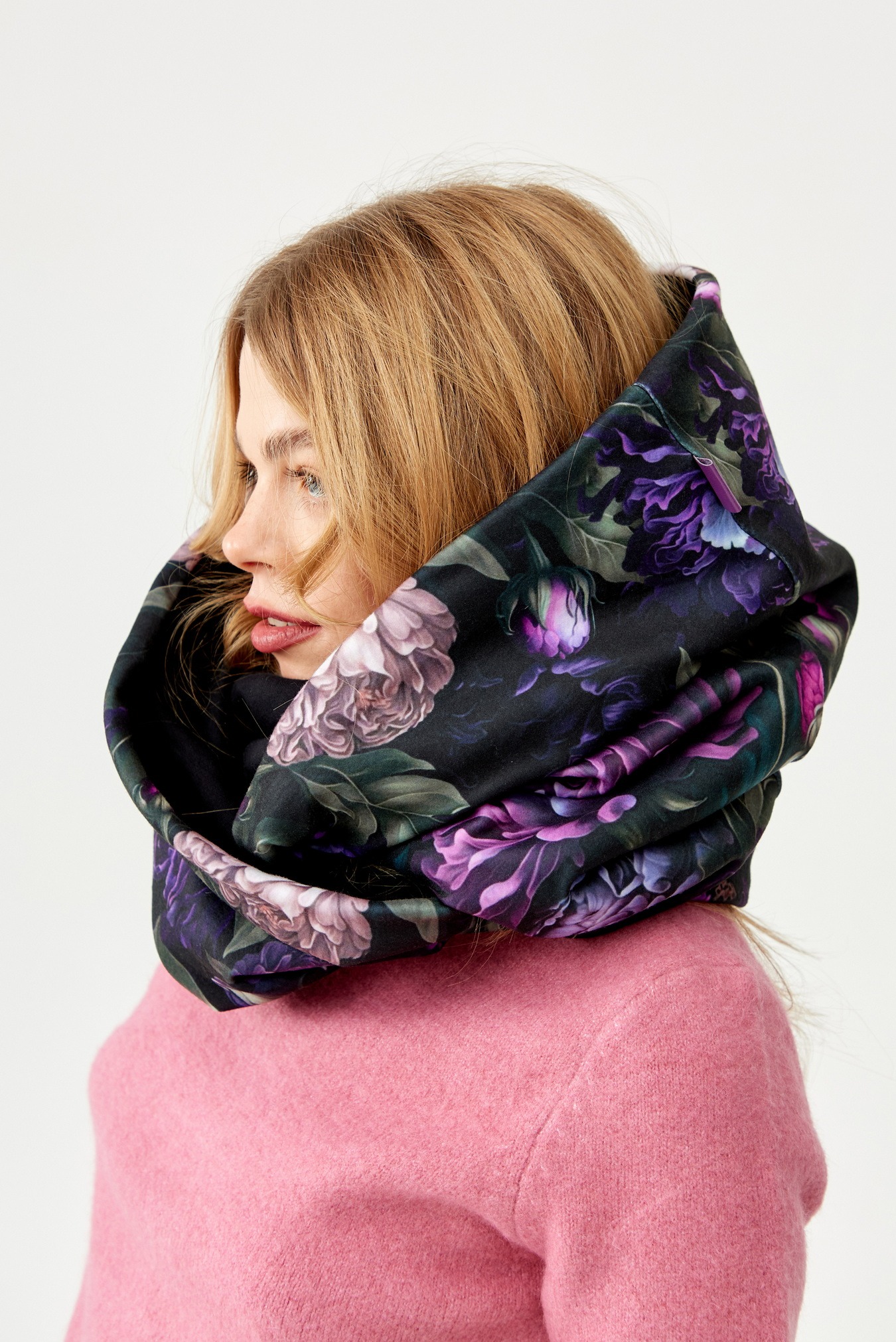 Profil kobiety z blond włosami, ubrana w różowy sweter i czarny komin z motywem fioletowych i różowych kwiatów na białym tle.