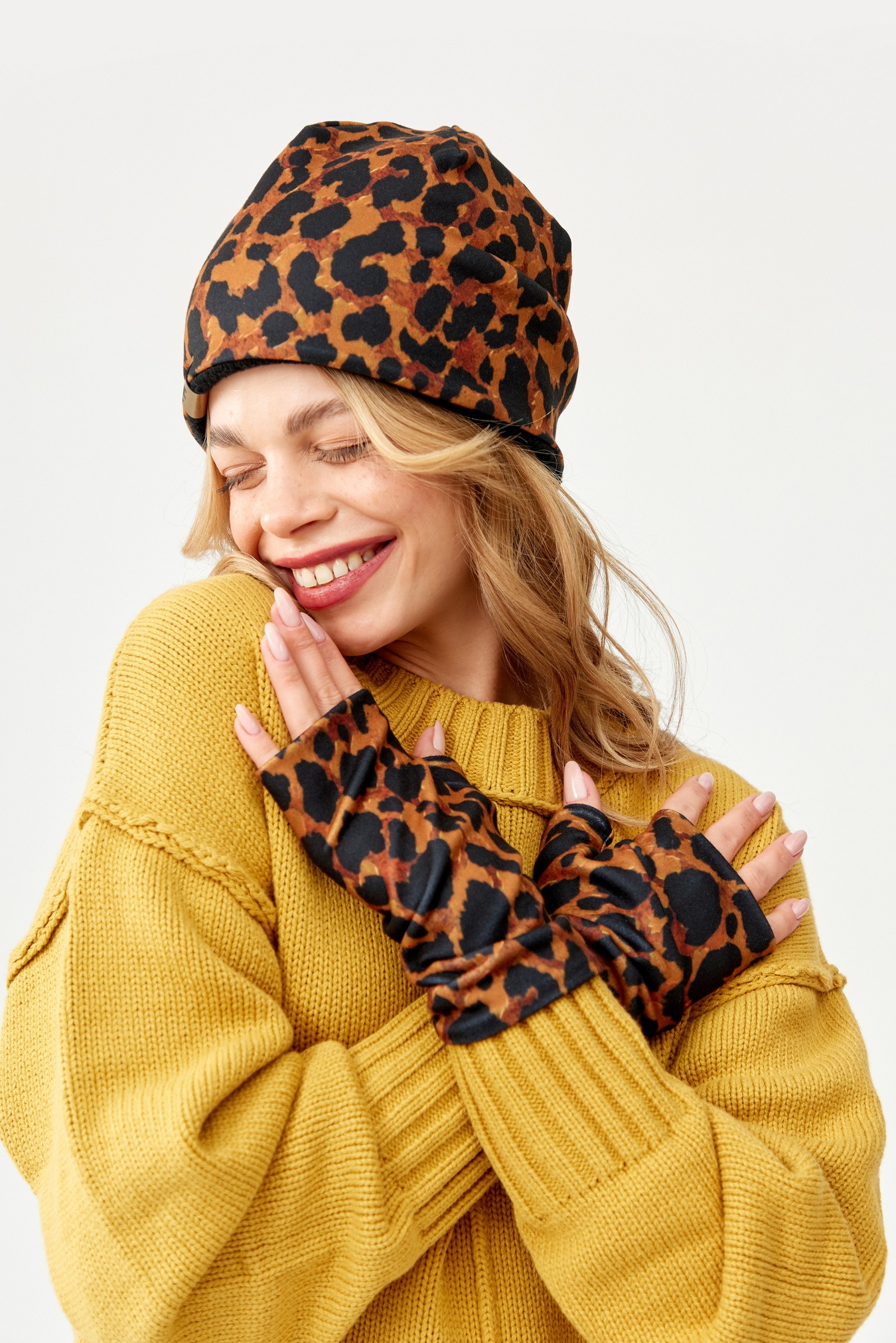 Uśmiechnięta blondynka w żółtym swetrze, czapce i rękawiczkach bez palców z motywem panterki, pozuje na białym tle.
