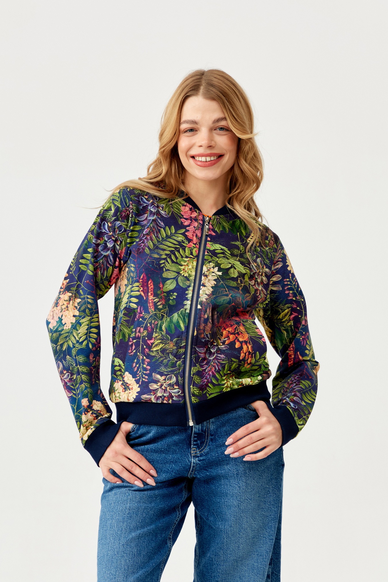 Uśmiechnięta blondynka w dżinsach i bomberce z motywem tropikalnych kwiatów i liści na granatowym tle, z rękami w kieszeniach.