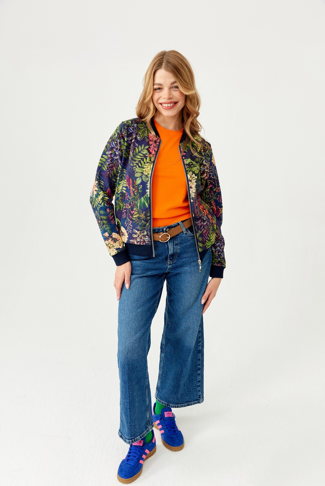Kobieta w dżinsach i jaskrawopomarańczowym swetrze, ubrana w bomberkę w kwiatowy wzór, pozuje na białym tle, eksponując stylizację.
