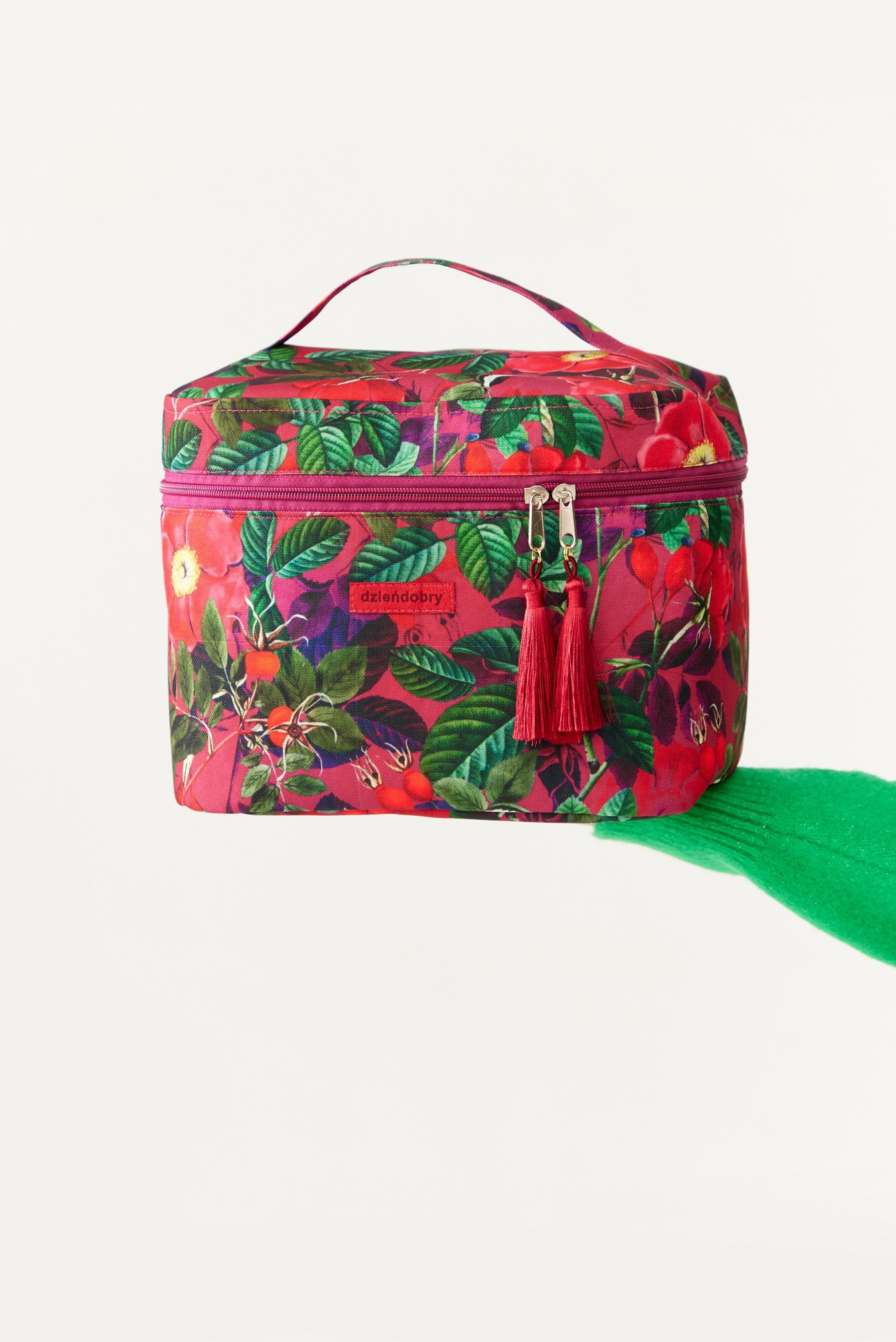 Pojemna kosmetyczka z uchwytem, zdobiona wzorem w róże i liście na różowym tle, z bordowym zamkiem błyskawicznym i ozdobnymi chwostami, leżąca na zielonej tkaninie.