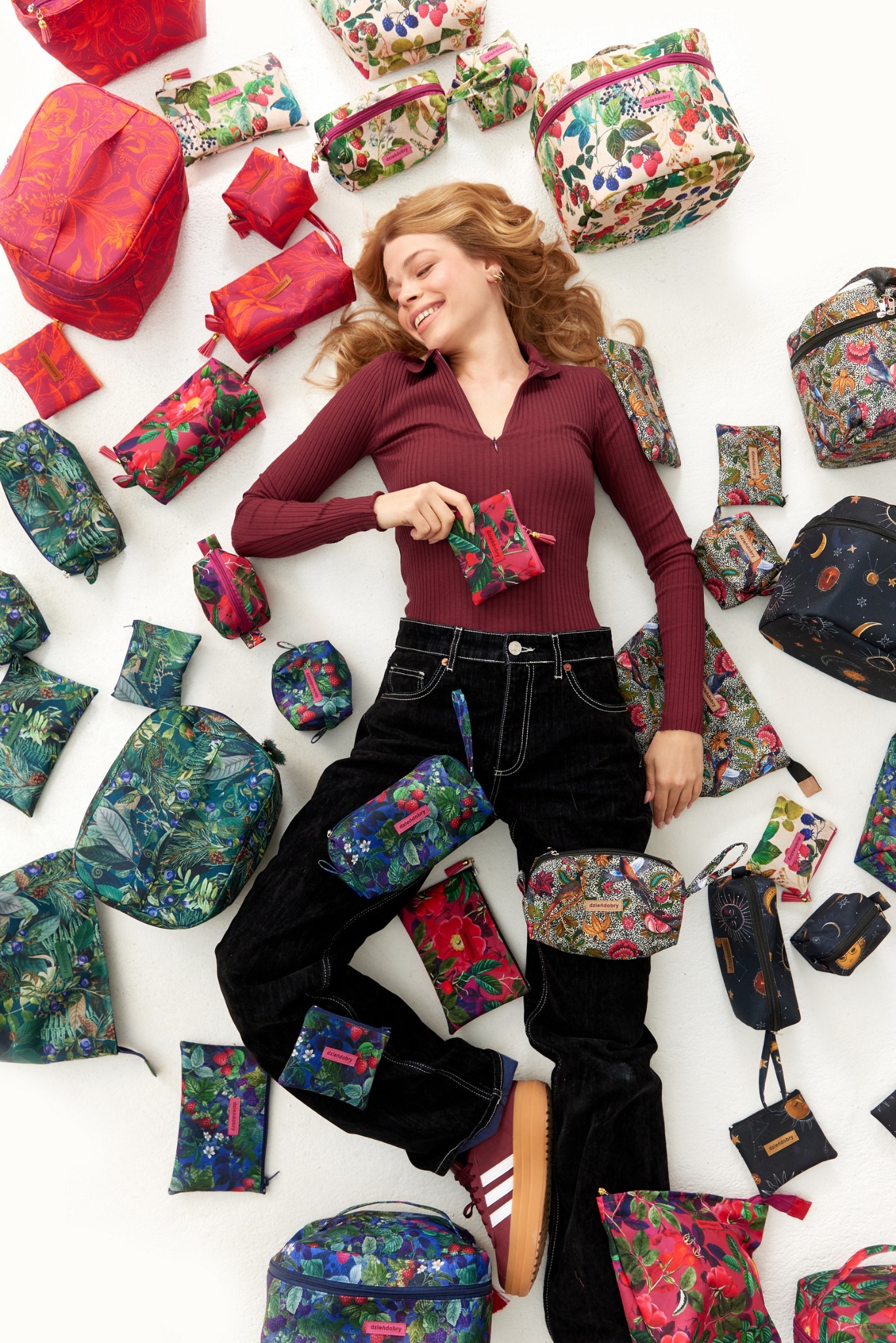 Uśmiechnięta kobieta leży na białym tle otoczona różnorodnymi kosmetyczkami i torbami z barwnymi, kwiatowymi i roślinnymi wzorami, prezentując małą kosmetyczkę w dłoni.