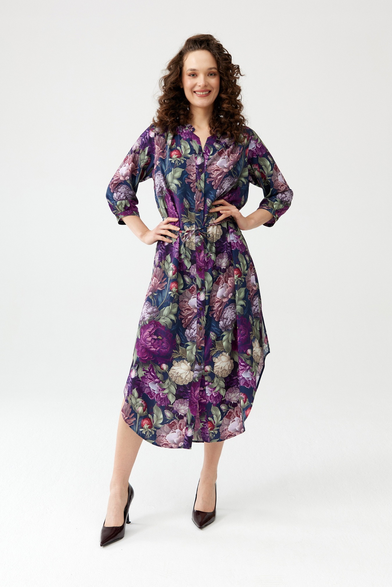 Kobieta pozuje w kwiecistej sukience koszulowej z rękawami 3/4 i lakierowanych szpilkach na białym tle.