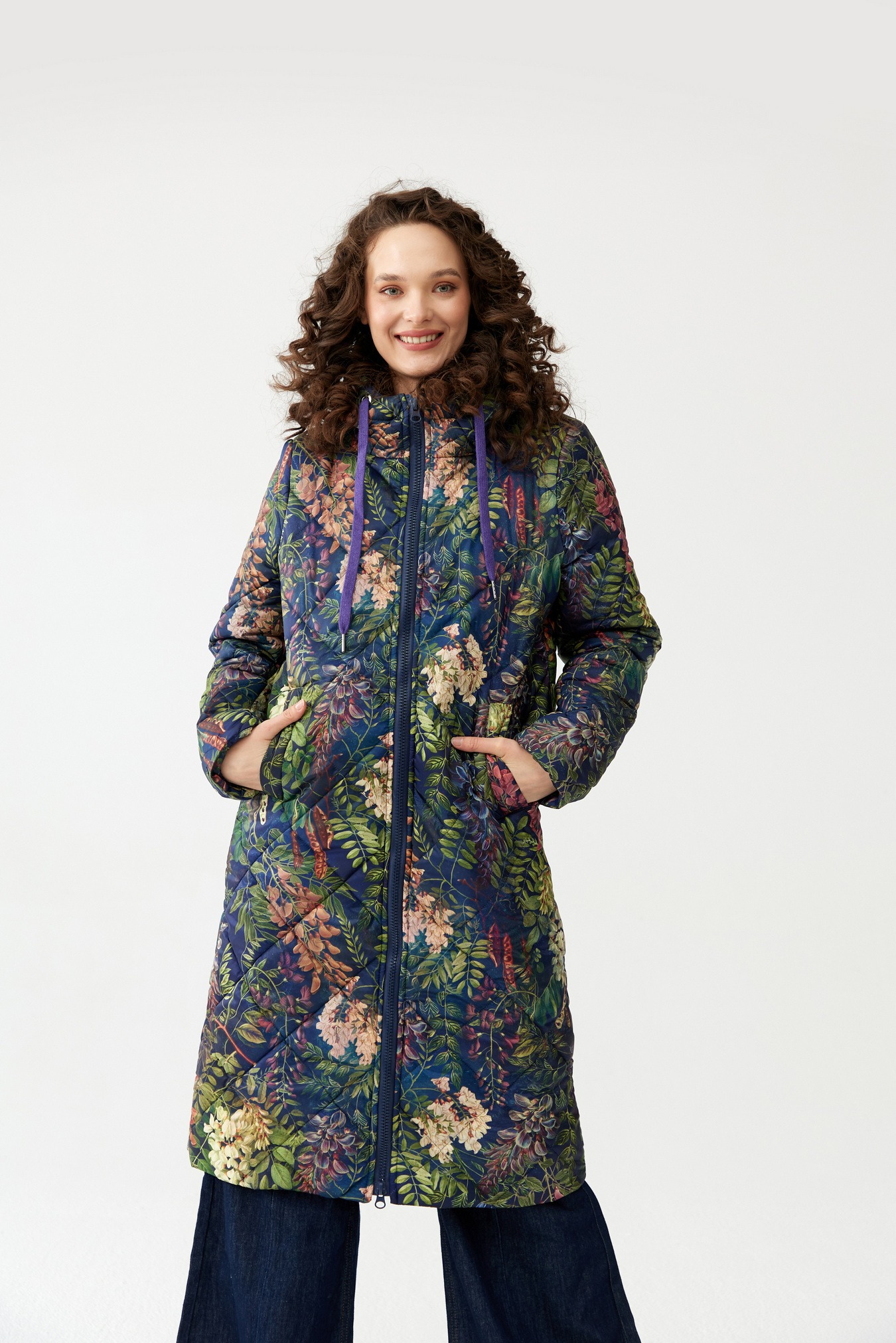 Kobieta z kręconymi włosami w długiej, pikowanej kurtce z wzorem w kwiaty i granatowymi sznurkami kaptura, ręce w kieszeniach, na białym tle.