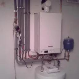 Biały piec gazowy Viessmann zamontowany nad białym zasobnikiem ciepłej wody, widoczne rury instalacji, dokumentacja techniczna na zasobniku, oświetlenie sufitowe.