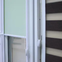 Białe okno PCV z roletami: gładka, pistacjowa roleta zwinięta częściowo i roleta dzień-noc w tonacji beżowo-brązowej. Widoczne detale mechanizmu łańcuszkowego.