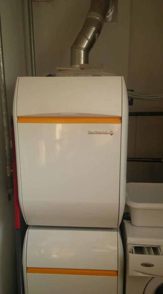 Biały, dwuczęściowy piec gazowy De Dietrich z żółtymi akcentami i rurą odprowadzającą spaliny, stojący obok pralki i pojemnika.