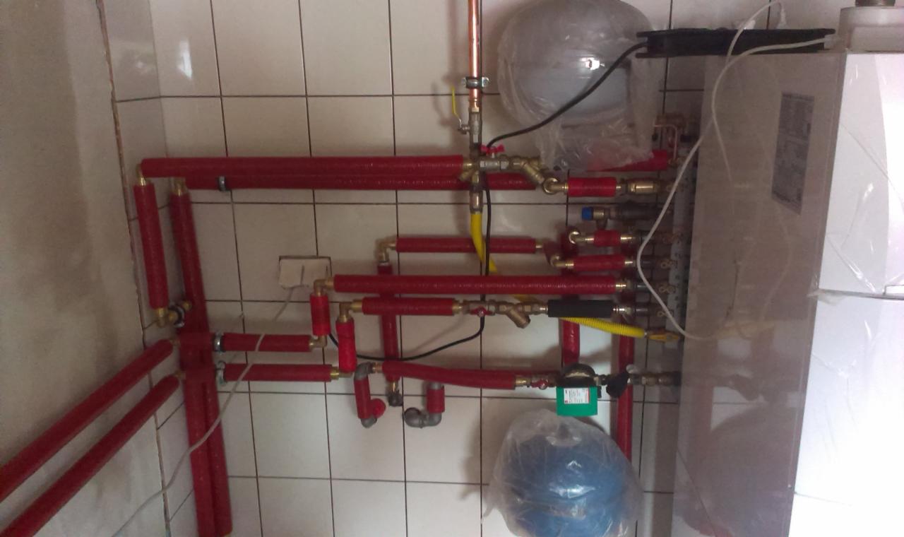 Złożona instalacja grzewcza z czerwonymi rurami i białym piecem na tle białych płytek. Widoczne liczne złączki i zawory. Elementy instalacji owinięte folią ochronną.
