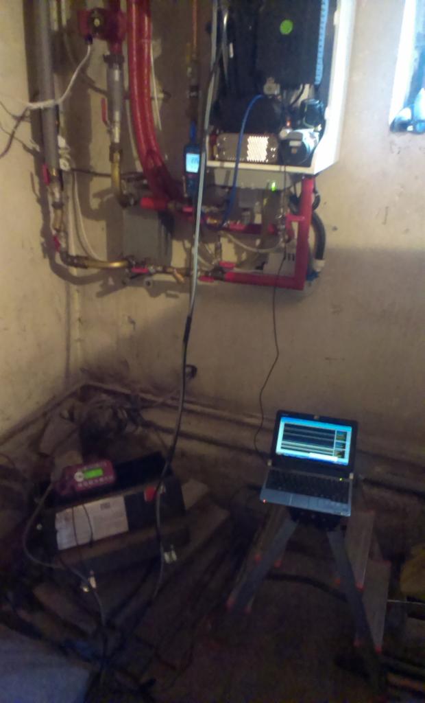 Piec gazowy z podłączonym laptopem i miernikiem podczas testowania. Widoczne rury i elementy instalacji w surowym pomieszczeniu.