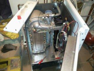 Otwarty piec gazowy w trakcie montażu, widoczne wnętrze z elementami instalacji, narzędzia obok. Kąt ujęcia z góry, oświetlenie mieszane.