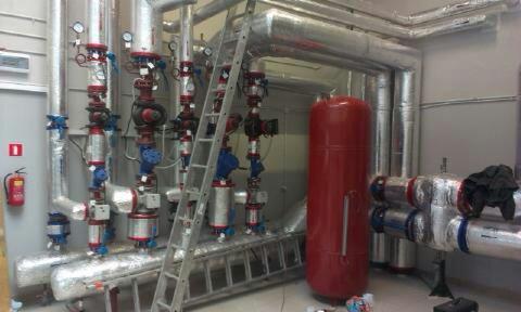 Kompleksowa instalacja grzewcza z izolowanymi rurami, zaworami i zbiornikiem wyrównawczym. Widoczna drabina i gaśnica. Realizacja w pomieszczeniu technicznym.