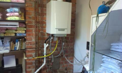 Zamontowany piec gazowy na ceglanej ścianie, widoczne rury i przyłącza. Półki z różnymi przedmiotami w tle. Instalacja wykonana schludnie, ale otoczenie nieuporządkowane.