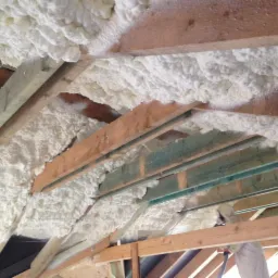 Ocieplenie poddasza pianką PUR, widoczne drewniane krokwie i biała, spieniona izolacja. Aplikacja pianki w trudno dostępnych miejscach.