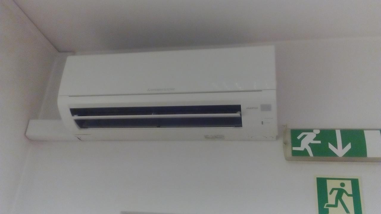 Biały klimatyzator Mitsubishi Electric zamontowany na jasnej ścianie obok zielonego znaku ewakuacyjnego z piktogramem biegnącej osoby i strzałką w dół.