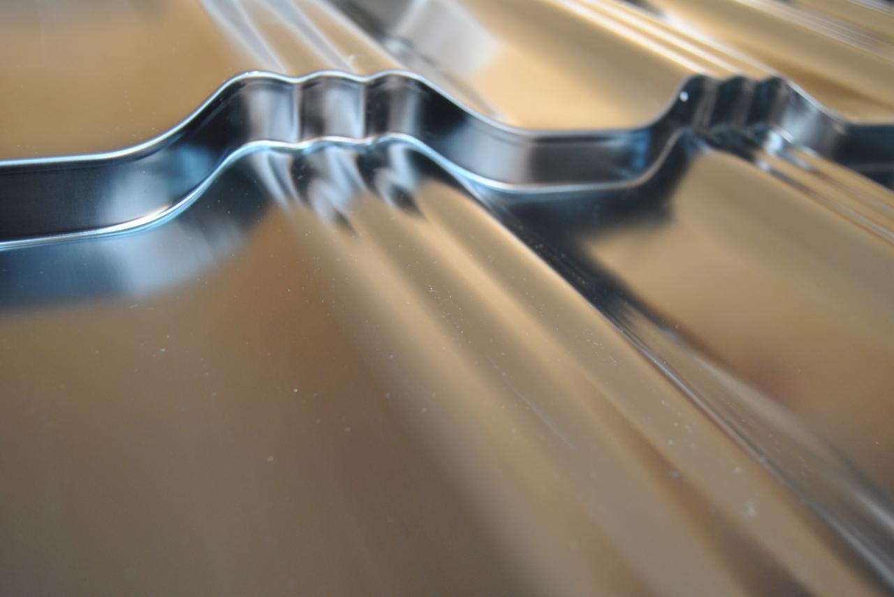 Błyszcząca blacha trapezowa w zbliżeniu, ukazująca regularne fale i metaliczny połysk. Widoczne delikatne refleksy światła podkreślają fakturę materiału.