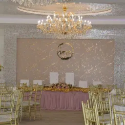 Elegancko udekorowany stół prezydialny z różowym obrusem, złotą girlandą i kwiatami, na tle ściany z dekoracyjnym wzorem i okrągłą ozdobą, oświetlony kryształowym żyrandolem.