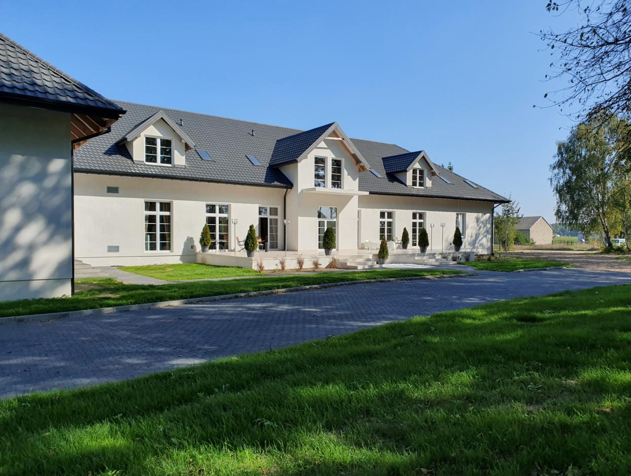 Elegancki budynek z białymi ścianami i ciemnoszarą dachówką, otoczony zadbanym trawnikiem i brukowanym podjazdem, z małymi drzewkami w donicach przed wejściem.