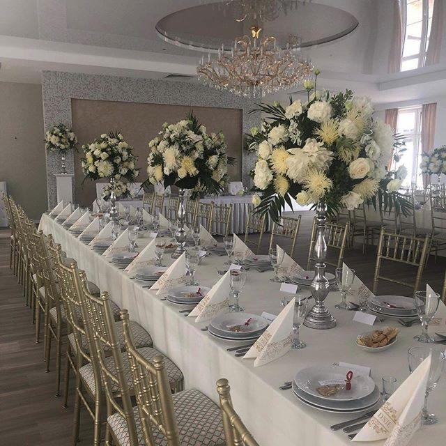 Uroczysta sala bankietowa z długim stołem nakrytym białym obrusem, dekoracje z białych kwiatów w srebrnych wazonach, złote krzesła, kryształowy żyrandol, elegancka zastawa i serwetki z subtelnym...