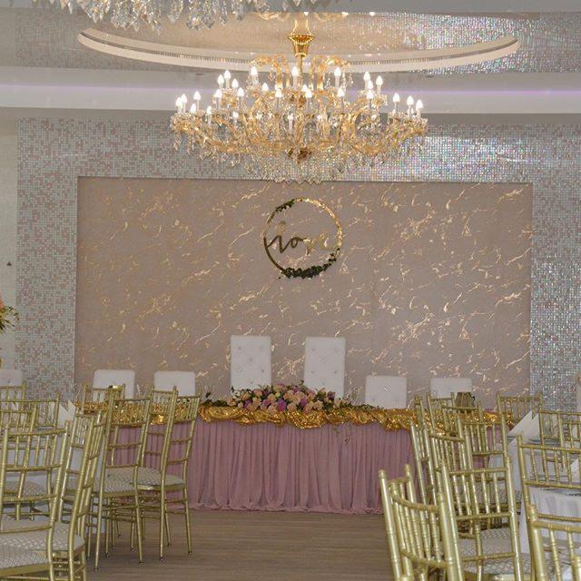 Elegancko udekorowany stół prezydialny z różowym obrusem, złotą girlandą i kwiatami, na tle ściany z dekoracyjnym wzorem i okrągłą ozdobą, oświetlony kryształowym żyrandolem.