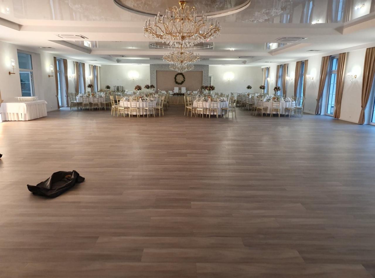 Ustawienie stołów z białymi obrusami i złotymi krzesłami w przestronnej sali balowej z parkietem, gotowej na przyjęcie, widoczne dekoracje kwiatowe na stołach i wieniec na ścianie