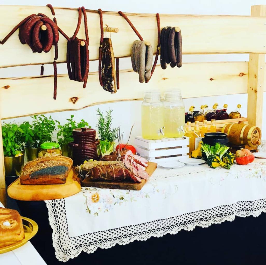 Stół z przekąskami: wędliny wiszące na drewnianej konstrukcji, chleb, pieczeń, lemoniada w słoikach, zioła w doniczkach, dekoracyjne dynie, całość na stole przykrytym białym obrusem z koronką.
