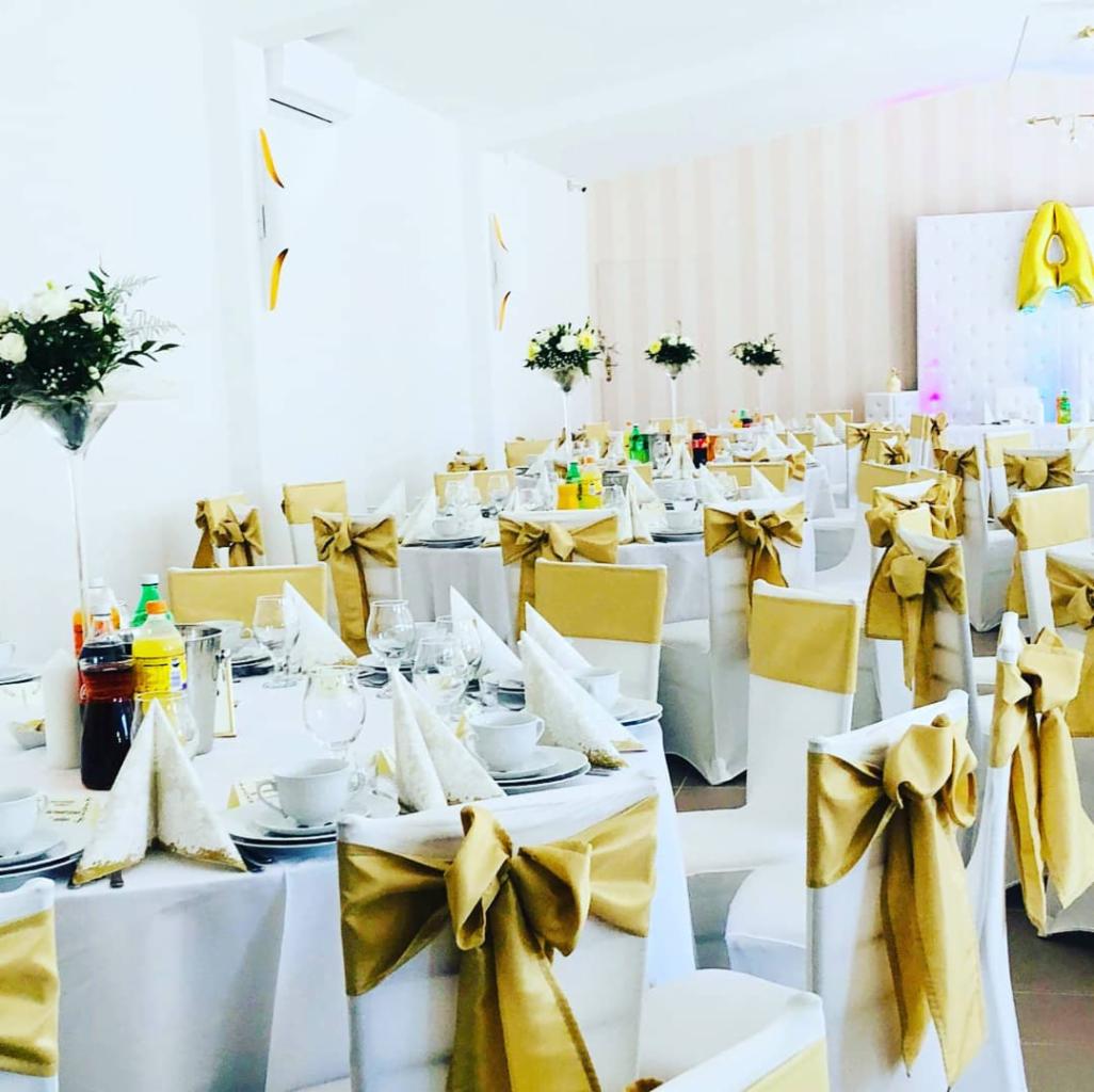 Sala bankietowa z białymi stołami i krzesłami udekorowanymi złotymi kokardami, nakrycia stołowe z białej porcelany, wysokie wazony z kwiatami, napoje, balon w kształcie litery A w tle.