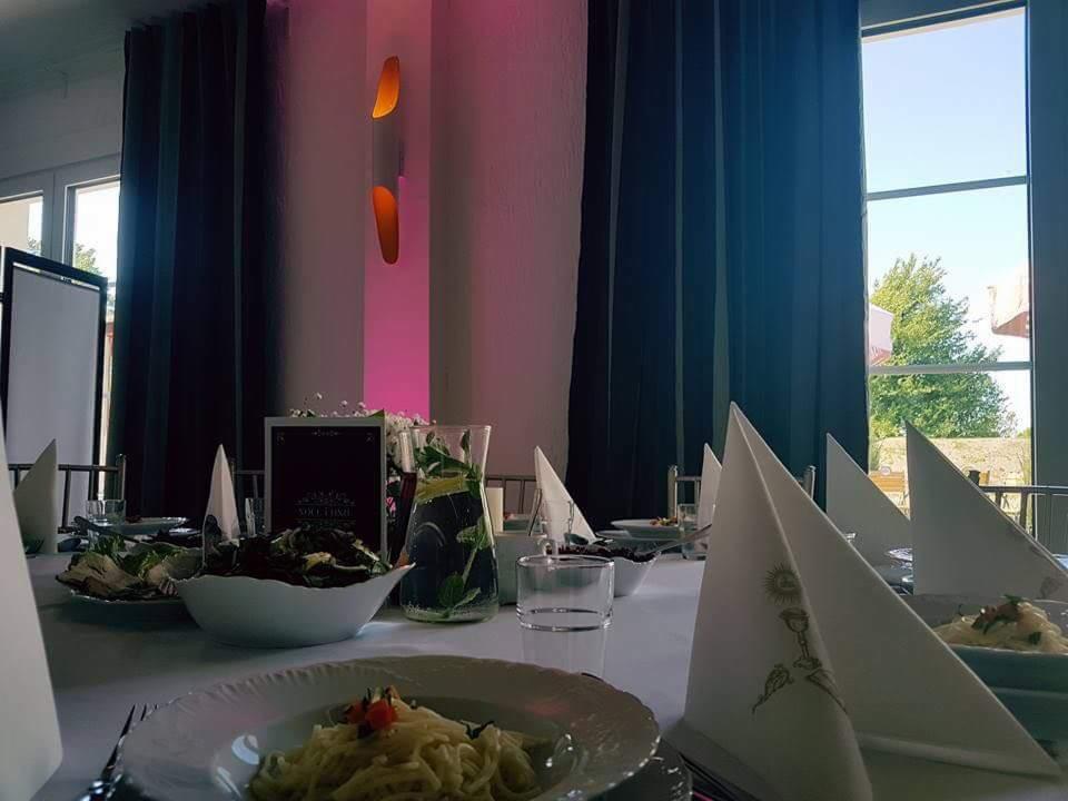 Uroczysty stół z dekoracjami i zastawą, ustawiony w sali z dużymi oknami i ciemnymi zasłonami, oświetlony ciepłym światłem.