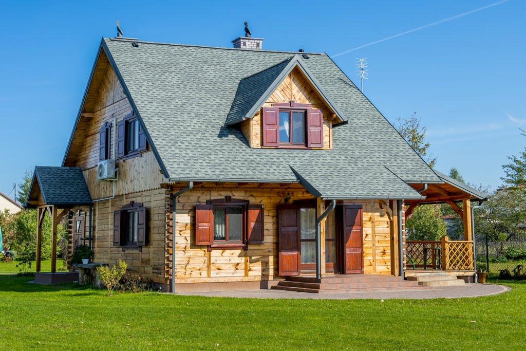 Jasny dom z bali z szarym dachem i brązowymi okiennicami, otoczony zielonym trawnikiem w słoneczny dzień. Widoczny klimatyzator na ścianie.