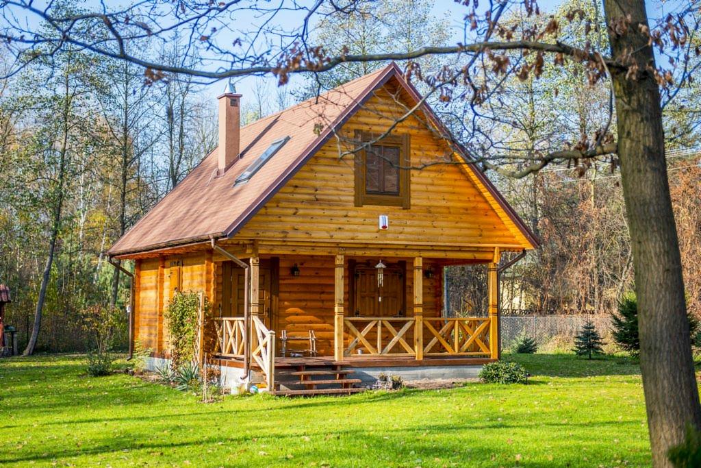 Urokliwy, parterowy dom z bali, z brązowym dachem i kominem, otoczony zielonym trawnikiem i drzewami. Widoczny fragment ganku z drewnianymi zdobieniami.