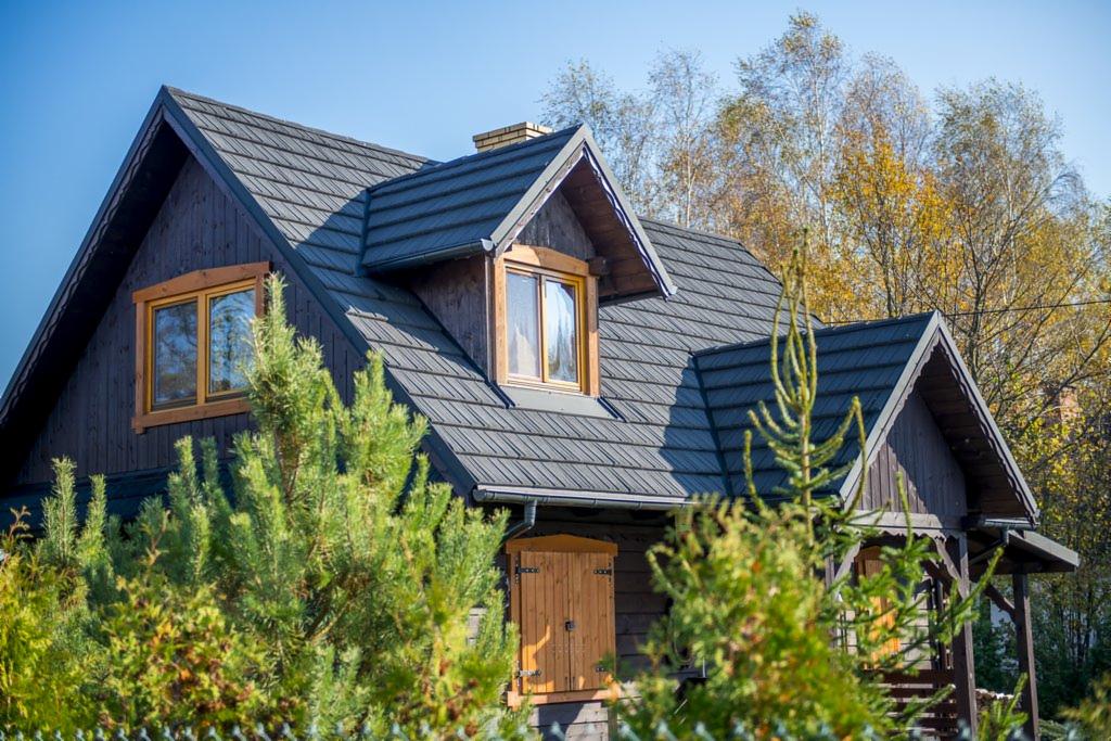 Ciemnobrązowy dom z drewnianą elewacją i dachem z szarej dachówki bitumicznej, częściowo zasłonięty zielonymi krzewami. Widoczne okna z drewnianymi ramami.