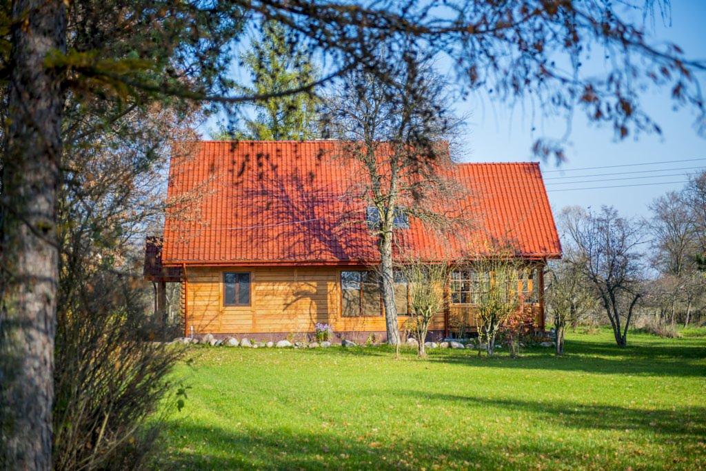 Drewniany dom z czerwoną dachówką w Warszawie, otoczony zielonym trawnikiem i drzewami, widziany w słoneczny dzień.