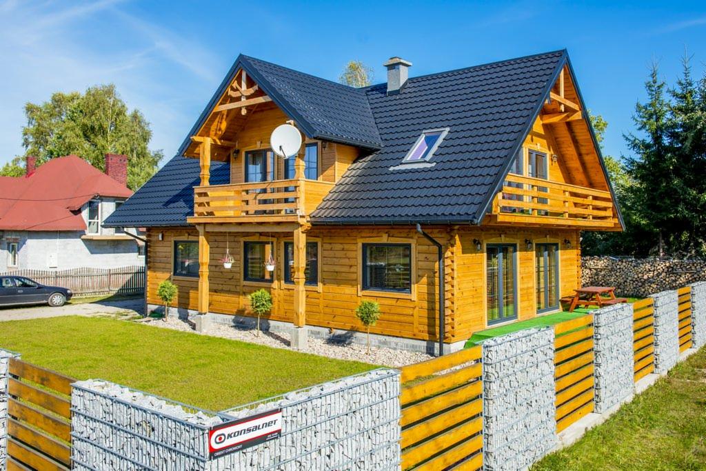 Drewniany dom z bali z balkonem i czarnym dachem, otoczony gabionowym ogrodzeniem. W tle widoczne drzewa i sąsiedni dom z czerwonym dachem.