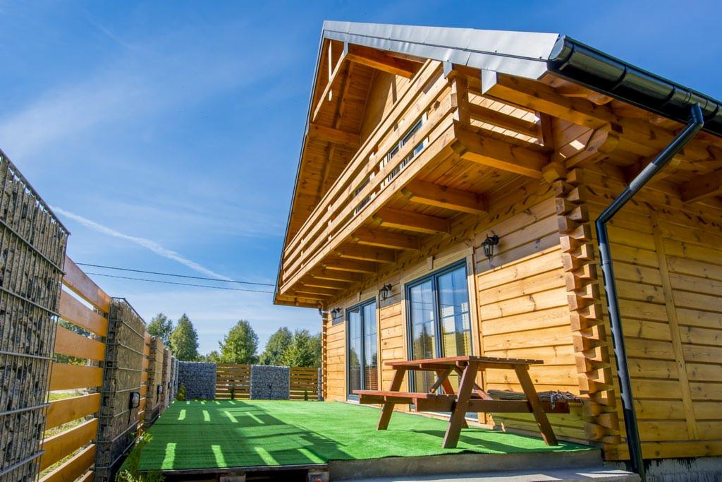 Drewniany dom z bali z balkonem i tarasem wyłożonym sztuczną trawą. Widoczny fragment ogrodzenia z gabionów. Słoneczny dzień, błękitne niebo.