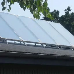 Cztery panele kolektorów słonecznych zamontowane na dachu budynku z szarą blachodachówką, widoczne liście drzew i fragment nieba.