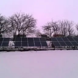 Rząd paneli fotowoltaicznych na śniegu, z drzewami w tle i wieżą telekomunikacyjną w oddali. Zimowy krajobraz z instalacją OZE.