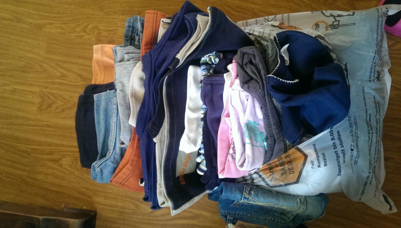 Stos ubrań dziecięcych i damskich, częściowo złożonych, częściowo w torbie z napisem 'Fair Kompetent Sozial', leżących na drewnianej podłodze. Różne kolory i fasony.