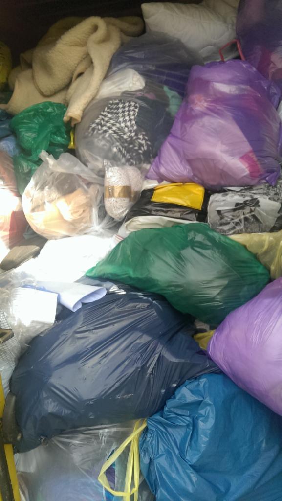 Pękate, kolorowe worki foliowe i stosy ubrań, prawdopodobnie odzieży używanej, wypełniające przestrzeń, częściowo w jasnych i przezroczystych workach.