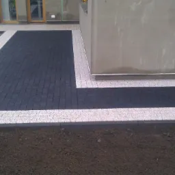 Ułożona nawierzchnia z kostki brukowej w dwóch kolorach: czarnym i jasnoszarym, z widocznym fragmentem budynku w tle i stertą kostki do ułożenia obok.