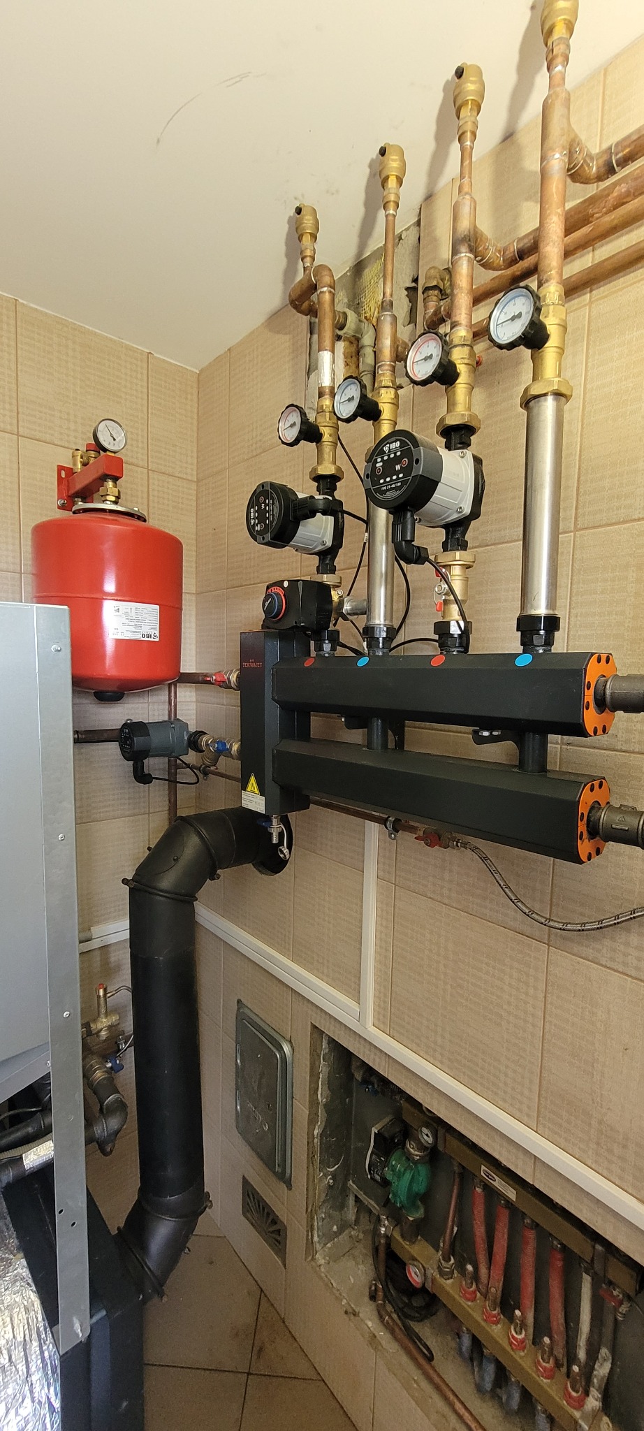 Złożona instalacja grzewcza z piecem, zbiornikiem wyrównawczym, pompami obiegowymi i rozdzielaczem, widoczne liczne rury miedziane i manometry.